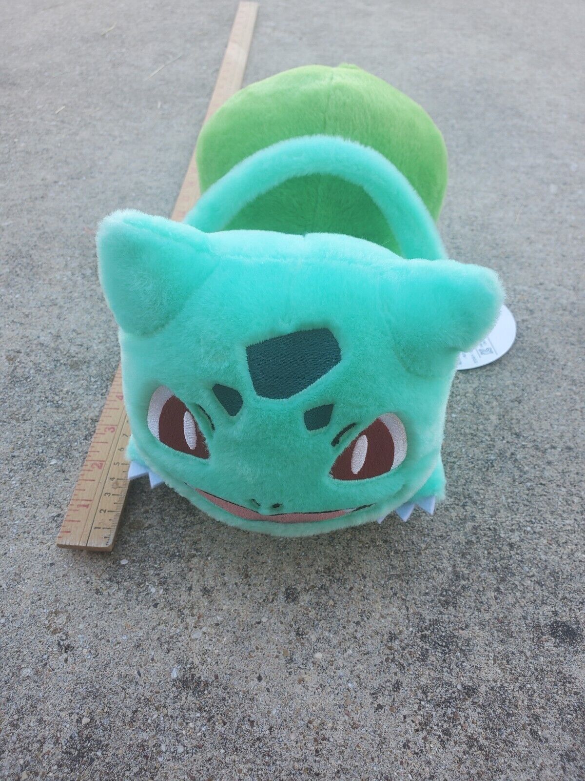 a plush of bulbasaur, a pokemon. bulbasaur is a small teal frog-like creature with dark teal spots on it's body, triangular red eyes, a wide smile, and a large green plant bulb growing on it's back. the plush has a zipper compartment in front of the bulb.