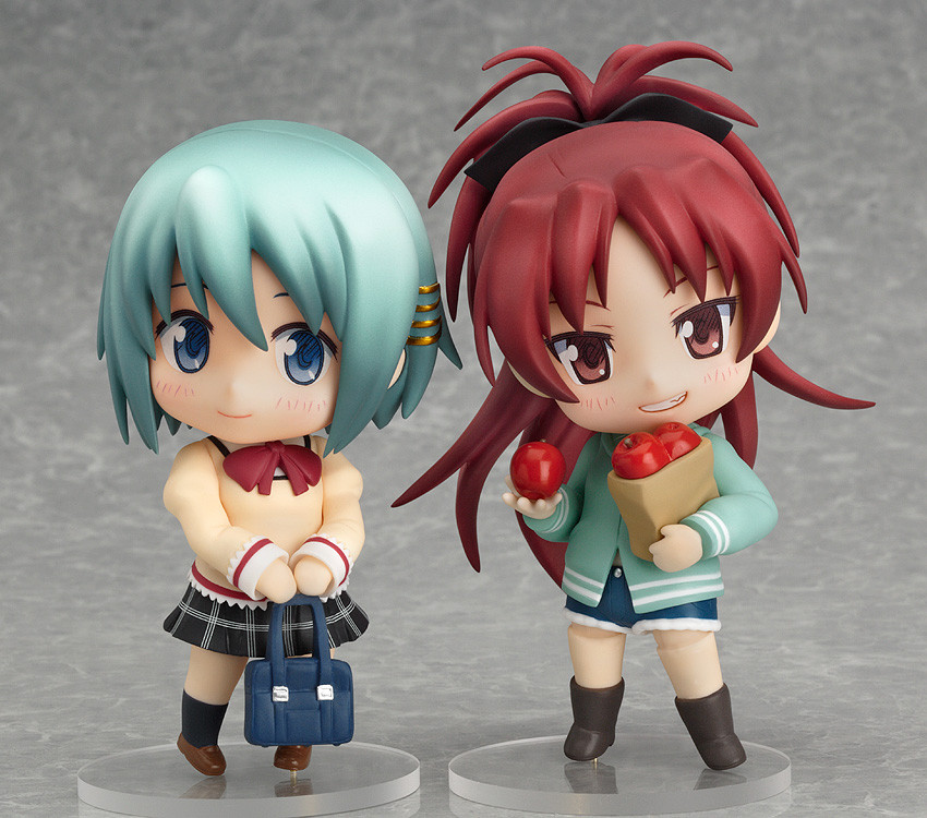 nendoroids of sayaka and kyouko, in their school uniform and casual clothes, respectively. sayaka is a taller girl with short blue hair, part of it on one side held back with gold hair pins, and blue eyes. sayaka's uniform consists of a cream blazer over a white shirt with back trim, a red bow at the neck, white trim on the edges of the sleeves and the bottom of the blazer, a black and white plaid skirt, black sports socks, and brown loafers. kyouko's casual outfit consists of a sea green hooded jacket over a black top, blue denim cutoff shorts, and brown boots. sayaka poses cutely, holding her schoolbag in front of her with both hands, looking shyly to her left. kyouko smiles confidently, holding a bag of apples in one arm and an apple in the hand of the other.