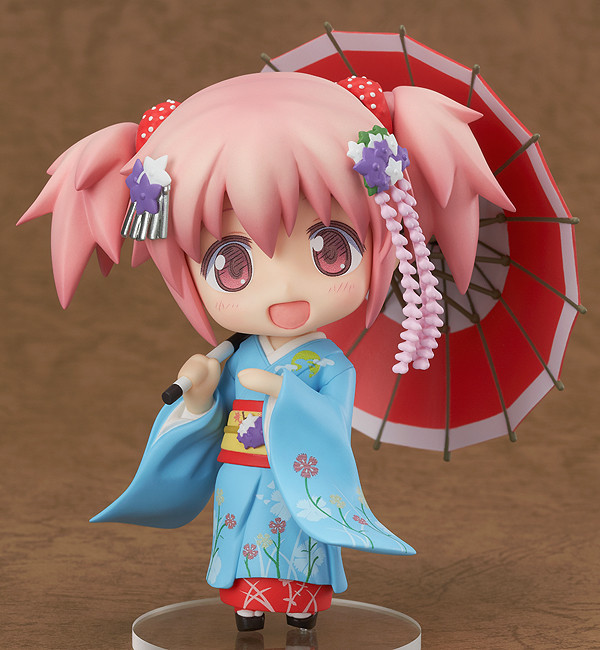 a nendoroid of madoka, a girl with pink hair tied into high pigtails that reach just past her chin, dark pink eyes, and fair skin. she's wearing a light blue kimono, adorned with a red and white floral pattern. on top of her kimono, she wears an orange and red darari obi, with a pocchiri made of white and purple flowers fastened to the front. the eri collar of her kimono is red and white, and the innermost layer of her kimono is red, and peeks out from the bottom of the rest of her kimono. she is wearing black geta with red straps on her feet, along with white socks. she has purple and white floral kanzashi and silver bira ogi fastened to her hair, and her hair is tied up in twintails with red arimachi kanoko, a tied silk hair ornament worn by maiko. she wears a bright smile, and is holding a red parasol in one hand.