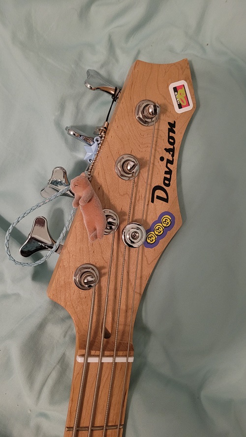 davison full size electric bass in black. the headstock has two small stickers on it, as well as two charms attached to the top tuning key: one of small, brown, sleepy cat, and a light blue one with a weaved loop and a star.