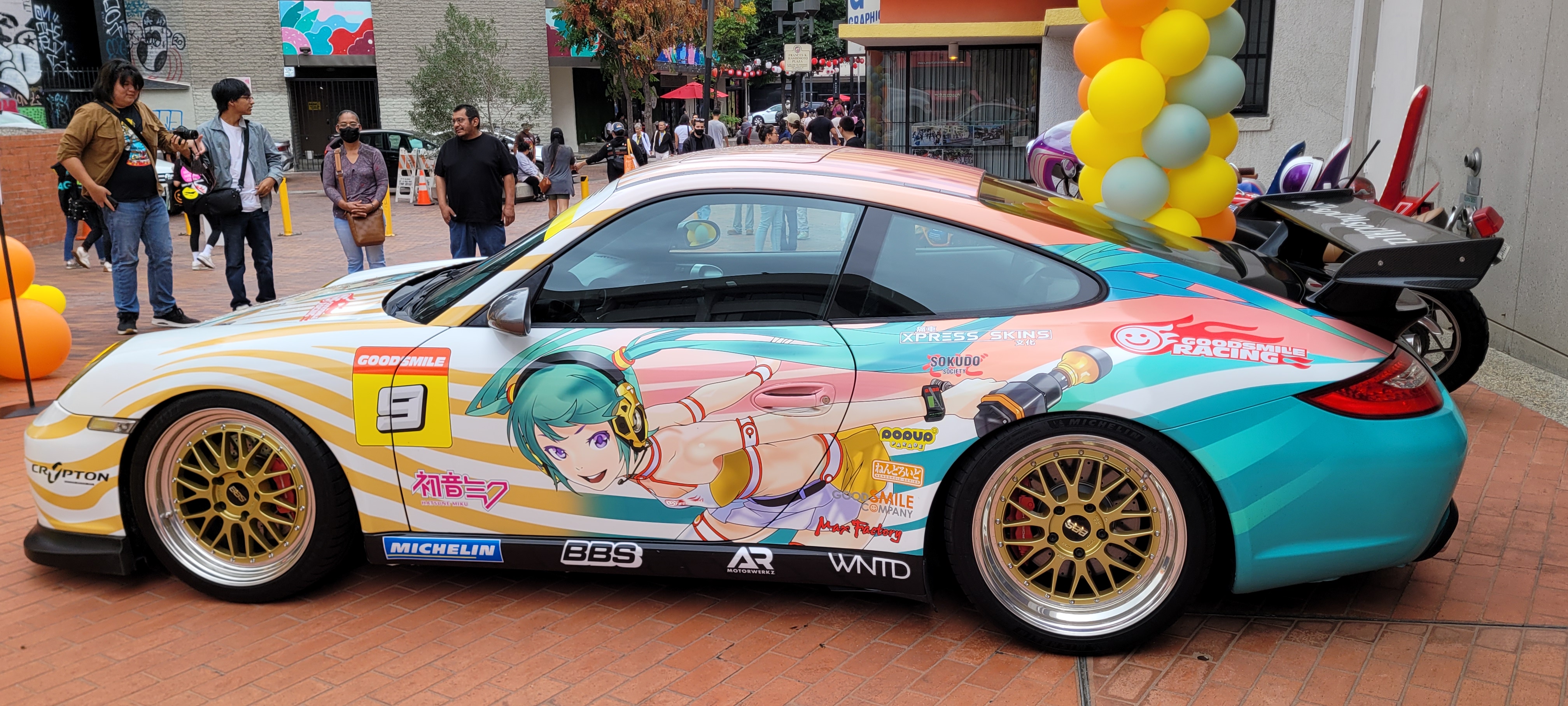 a photo of the miku itasha car. the car is primarily orange and yellow near the front, with the colors gradiating to blue near the back of the car. an ilustration of miku in an orange racing uniform holding an orange impact wrench is featured prominently, alongside several sponsor logos.