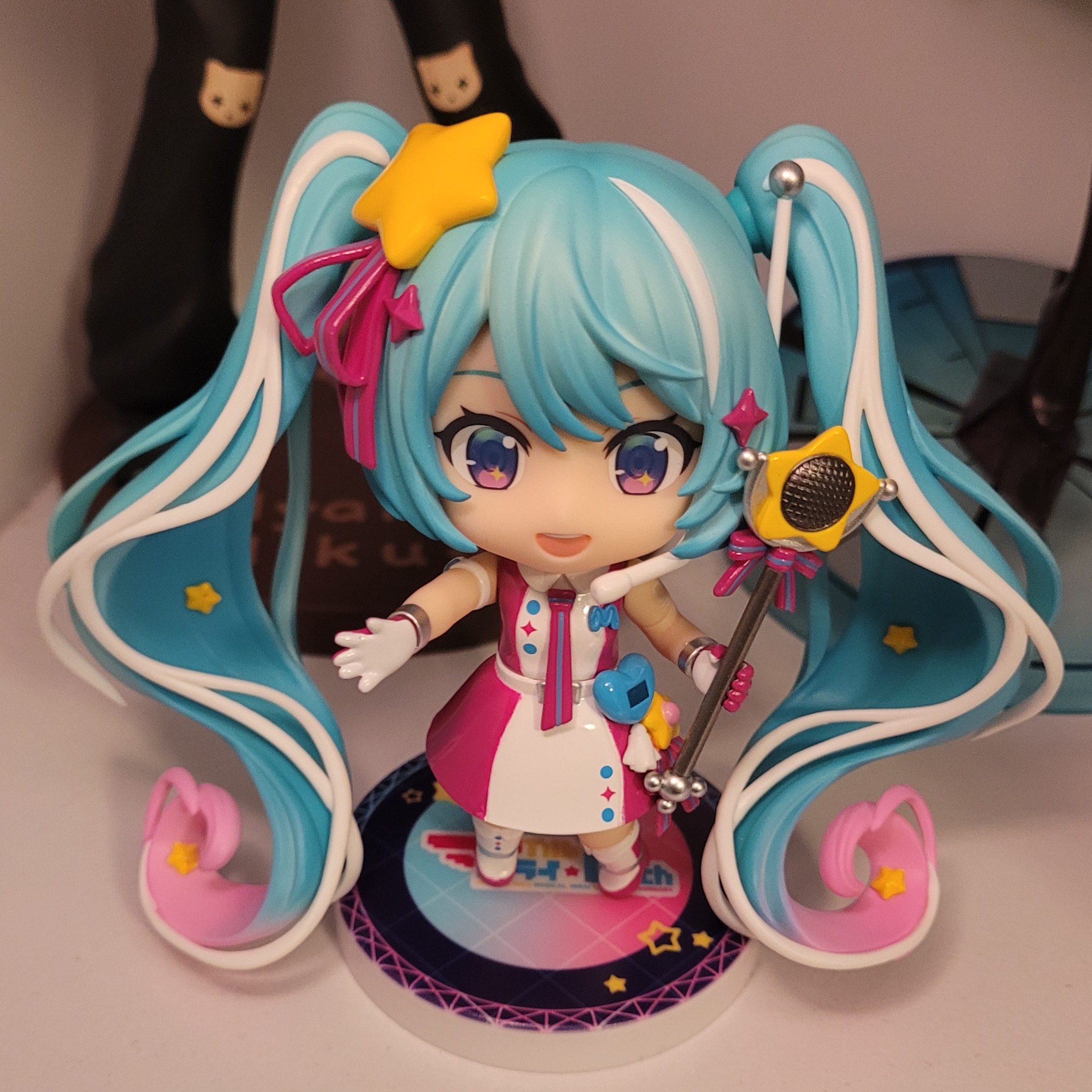 a photo of the Magical Mirai 10th Anniversary Miku Nendoroid. She wears a magenta and white dress with a magenta and white tie as well as a blue heart, yellow star, and magenta bow attached to her hip on her belt. She has long blue twintails that turn pink near the bottom and also have white streaks. She wears a large yellow star with a magenta bow attached on her left pigtail. She is also wearing a white headset that has a long white antenna with a silver top on the right side of her head. She's holding a star baton with a magenta bow.