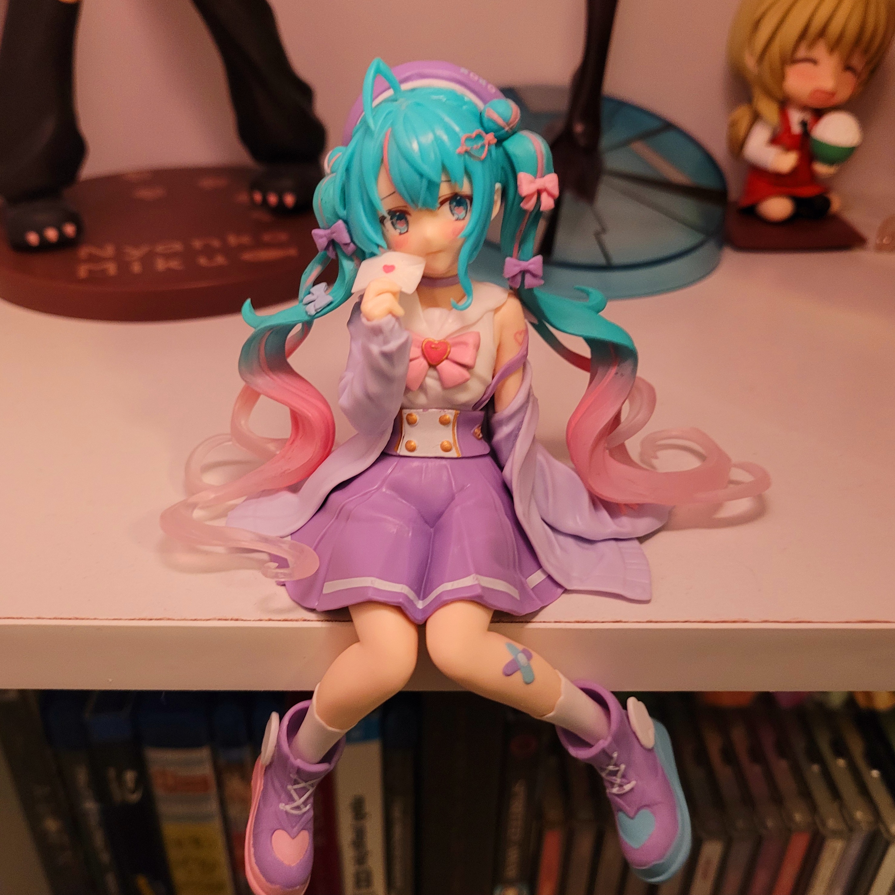 a photo of a miku figure. she is sitting, holding a small envelope with a pink heart on it. her outfit is primarily purple, featuring a purple skirt with suspenders, white shirt with a pink bow with a heart on it, and a light purple sweater. she also wears a purple beret. her pigtails have small hair buns tied near the top, and her pigtails fade from light blue to pink near the ends. there are also a few small purple and pink hair bows in each pigtail. she wears purple high top shoes with small white wings on the outer side of each, with the heart on the toe of the left shoe being pink and the heart on the toe of the right shoe being blue, as well as white socks with scalopped edges. she has a set of purple and blue bandaids on one of her legs.